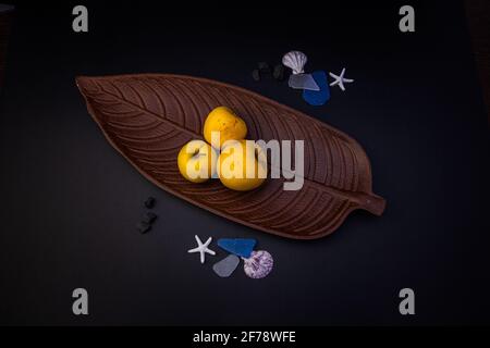 Three yellow apples placed on a brown leaf-shaped plate with a few shells, on a black background Stock Photo