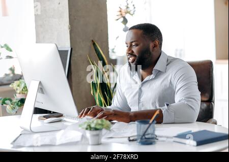 Handsome influential confident African American business leader, broker, manager or lawyer, working in an office at a computer, communicating online with a colleague or client, typing on the keyboard Stock Photo