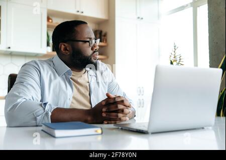 Successful handsome african american bearded man with glasses, freelancer or employee, works at home uses laptop, looks thoughtfully aside, thinks about business project, hands folded in front of him Stock Photo