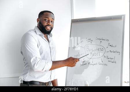 Teaching and learning concept. Smart successful african american bearded stylish business coach stands near a whiteboard, looks at the camera, smiles friendly Stock Photo