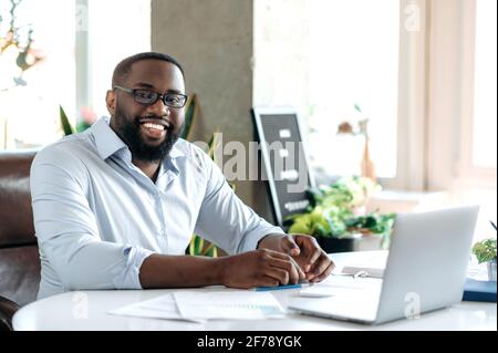 Successful handsome confident bearded african american businessman, real estate agent or ceo dressed in formal stylish clothes and glasses, sits at workplace, looking at camera, smiling friendly Stock Photo