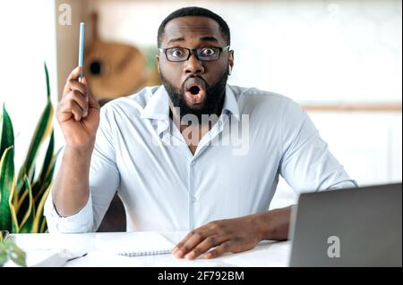 Portrait of excited shocked bearded african american man with glasses in formal shirt, sitting at table and showing idea gesture, looking directly at camera with surprised face expression Stock Photo