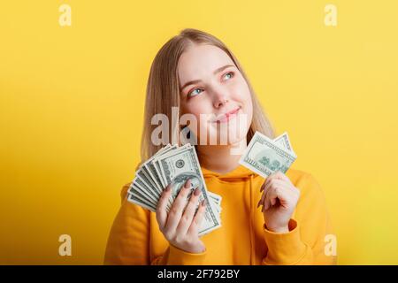 Happy teenage blonde girl holding in hands money cash dollars dreaming thoughtfully isolated on color yellow background. Portrait young excited smilin Stock Photo