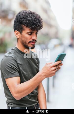 vertical portrait of a latin american young man checking his smart phone in the street. He is wearing casual summer clothes Stock Photo