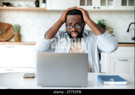 Shocked African American bearded man with glasses sits at his desk at home, looks at his laptop in confusion, reads bad news, received an unexpected message, failed deal, holds head with hands Stock Photo