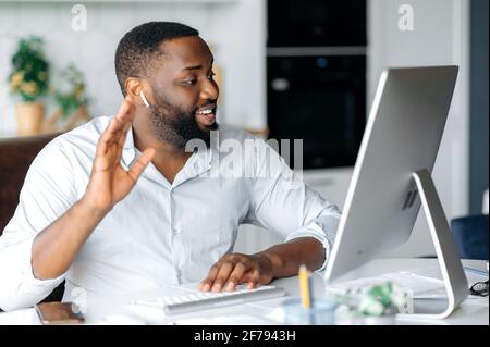 Attractive friendly successful young african american businessman, broker, or lawyer working remotely at computer, talking to employee or client by online conference, greeting with hand gesture, smile Stock Photo