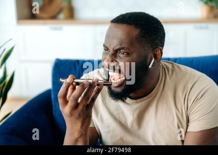 Outraged angry outraged African American guy, sitting on a sofa in the living room, uses wireless earphones, shouting while talking on speakerphone by cellphone, irritatedly recording an audio message Stock Photo