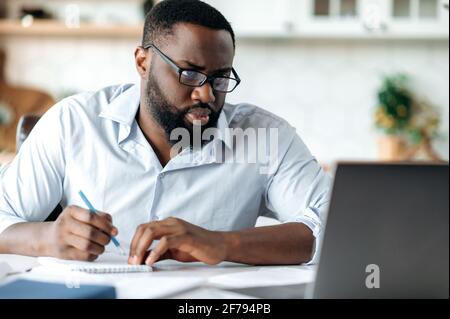 Serious purposeful intelligent african american man with beard wearing glasses, lawyer, broker or business leader, works remotely, sits at desk, takes notes, looks with concentration at laptop Stock Photo