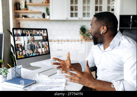 Successful stylishly dressed African American business man sits at work desk at home, looks at the computer screen, communicates on a video conference with his colleagues, discusses a business plan Stock Photo