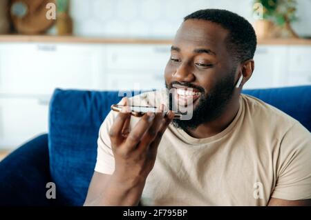 Close-up of handsome friendly confident african american guy sitting in living room on sofa talking or recording audio message on smartphone for friend or colleague, smiling happily Stock Photo