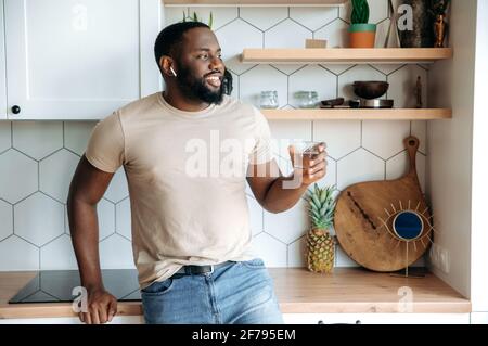 Happy attractive healthy African American bearded man, stands at kitchen, in casual clothes, with a glass of clean water in his hand, looks to the side, smiles friendly. Healthy lifestyle concept Stock Photo