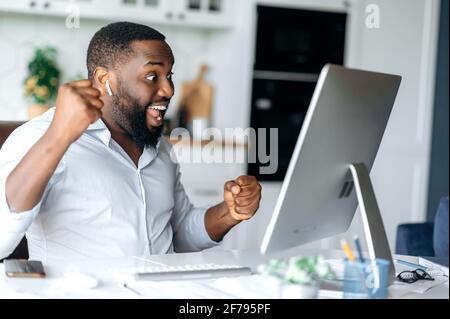 Successful cheerful confident african american male entrepreneur, freelancer or lawyer dressed in stylish clothes, working home, celebrating success, victory or good profit, gesturing with his hands Stock Photo