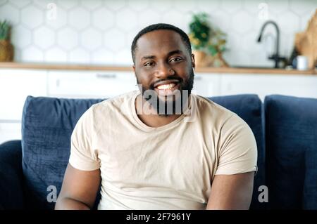 Close-up portrait of handsome happy young confident african american bearded man, sitting on sofa at home in living room, wearing t-shirt, looking at camera and smiling friendly Stock Photo