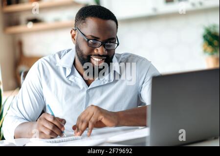 Friendly successful confident african american bearded male with glasses, broker, lawyer or manager working remotely, taking notes, talks by conference call with colleagues, distant learning, smiling Stock Photo