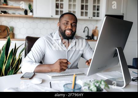 Portrait of a confident successful African American businessman, real estate agent, lawyer or manager, wearing a stylish shirt, working remotely at the computer, looking at camera, smiling friendly Stock Photo