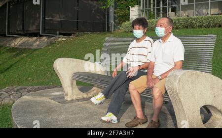 Senior Asian couple wearing face mask sitting on a bench in a park. Going outdoors during Covid-19 pandemic concept. Stock Photo