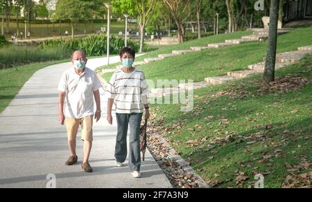 Senior Asian couple wearing face mask taking a walk in a green park. Healthy lifestyle during pandemic concept. Stock Photo