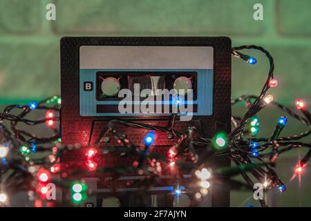 An audio cassette in a black plastic case is reflected on a black matte surface. Colorful lights are garlanded around it. Close-up Stock Photo