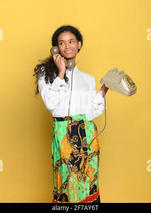 Trendy young Dominican woman in brightly colored skirt and smart white blouse standing talking on a vintage telephone over a yellow studio background Stock Photo