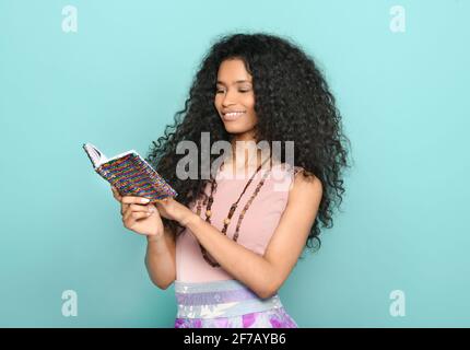 Happy young Dominican woman with lovely long curly hair standing reading notes from a colorful notebook with a beaming smile over a turquoise studio b Stock Photo