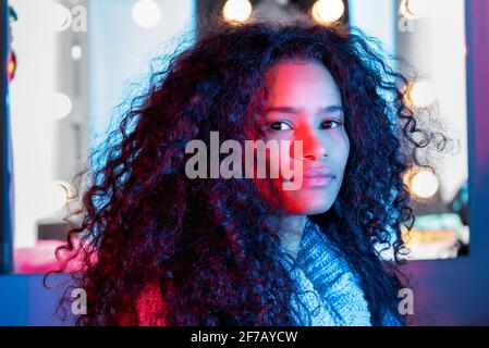 Cute attractive black woman frame by glowing lights with thick curly hair watching the camera with a sideways glance side lit by colorful pink studio Stock Photo