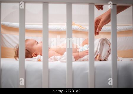 A mother puts the baby to sleep in the crib. Close-up. Side view, through the bars of the cradle. Stock Photo