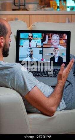 Tired man sitting comfortable on sofa while chatting with teammates about social communication during business videocall conference using laptop computer late at night in kitchen Stock Photo
