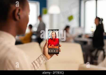 Over the sholder of black employee discussing with partner on video call using phone in start up business office. Black woman during video meeting, online conferencel planning new financial project Stock Photo