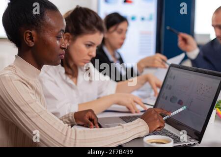 Cheerful african business lady typing on laptop and smiling sitting at desk in busy start up office. African woman using laptop in meeting broadroom, negotiations speaking with clients Stock Photo