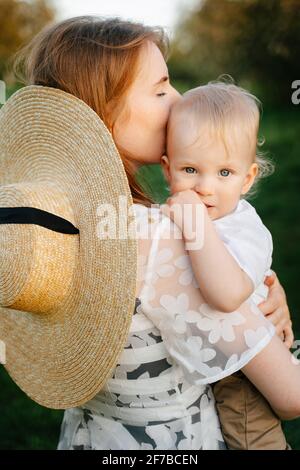 A loving young mother holds the baby boy in her arms hugging him, the child looks into the camera. Stock Photo
