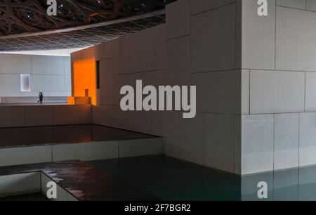 Two men walking in the direction of a white wall changed to orange due to the setting sun. Louvre museum. Abu Dhabi, United Arab Emirates. Stock Photo
