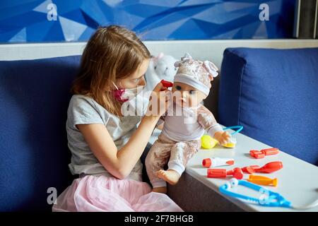 A little girl in medical mask sits on the sofa, plays a doctor, treats to the doll Stock Photo