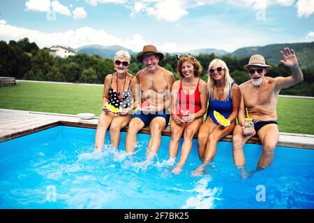 Group of cheerful seniors sitting by swimming pool outdoors in backyard, looking at camera. Stock Photo