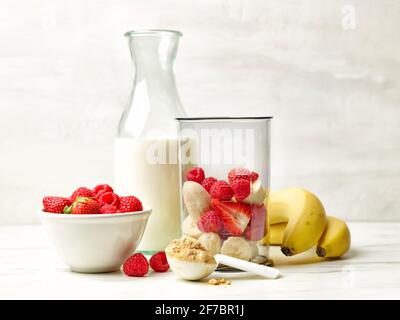 fresh banana pieces and red berries in plastic transparent blender container and milk bottle on kitchen table ready for making healthy breakfast smoot