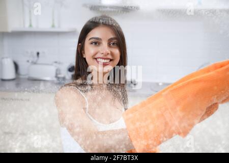 Close up of smiling cute woman washing glass with rag in kitchen. Concept of house cleaning. Stock Photo