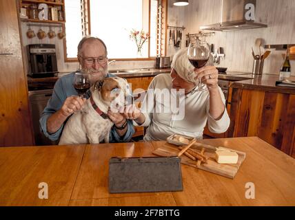 Happy senior couple with pet dog and wine toasting online video calling friends and family celebrating winter holidays remotely. Coronavirus second wa Stock Photo
