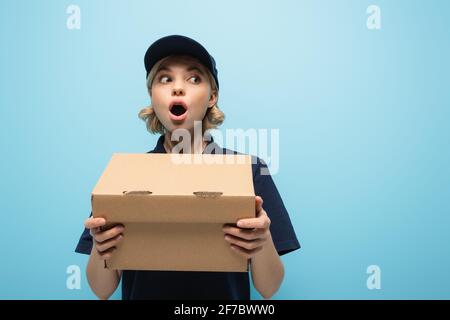 shocked courier looking away while holding parcel isolated on blue Stock Photo