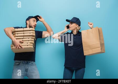 shocked muslim delivery man looking at colleague accepting order on smartphone on blue Stock Photo