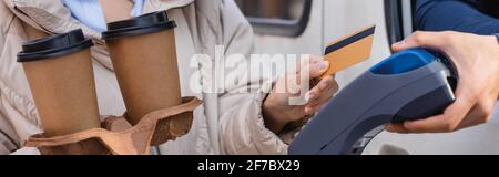 partial view of delivery man holding payment terminal near woman with paper cups and credit card, banner Stock Photo