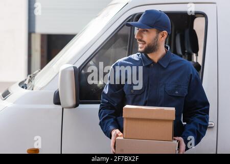 smiling arabian courier looking away while holding parcels near car Stock Photo