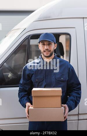 cheerful muslim postman holding parcels while standing near car outdoors Stock Photo