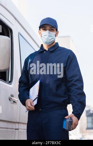 arabian postman in protective mask standing near car with payment terminal and clipboard Stock Photo