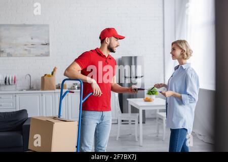 muslim postman giving clipboard to woman while standing near trolley with parcels on blurred foreground Stock Photo