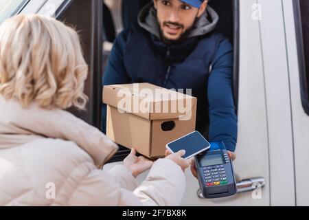 arabian delivery man giving payment terminal and parcel to woman with cellphone on blurred foreground Stock Photo