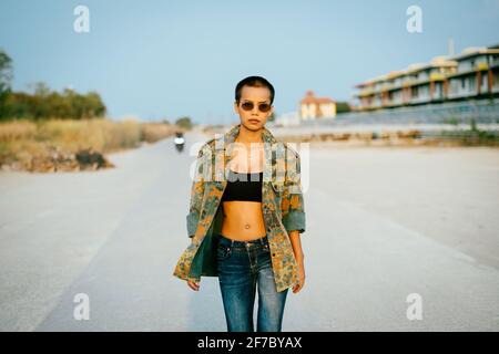 Young short hair Asian woman with sunglasses wearing sports bra and army jacket, walking straight towards the camera with confidence and determination Stock Photo