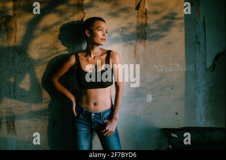 Young short hair Asian woman wearing sports bra and blue jeans standing against a wall with graffiti and shadows, looking to the side into the light. Stock Photo