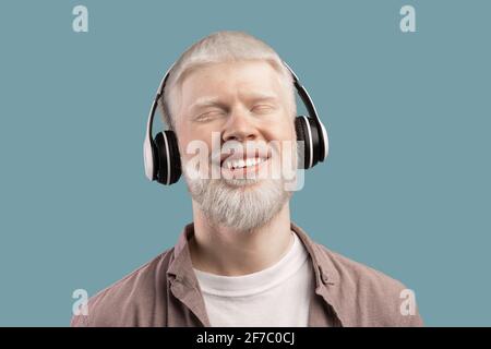 Music lover. Peaceful albino man in wireless headphones listening audio on turquoise studio background Stock Photo