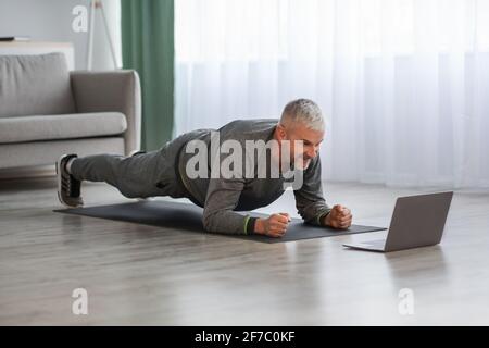 Athletic mature bearded man doing exercises at home, using laptop