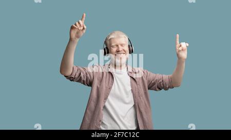 Happy albino man enjoying music in wireless headphones, dancing and raising fingers up over turquoise studio background Stock Photo