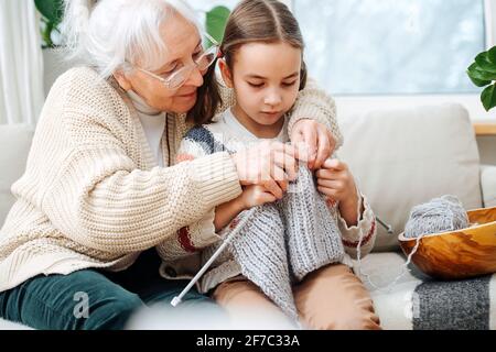 Skillful granny sitting on a couch with her granddaughter She's teaching her how to knit, holding hands over hers. Stock Photo
